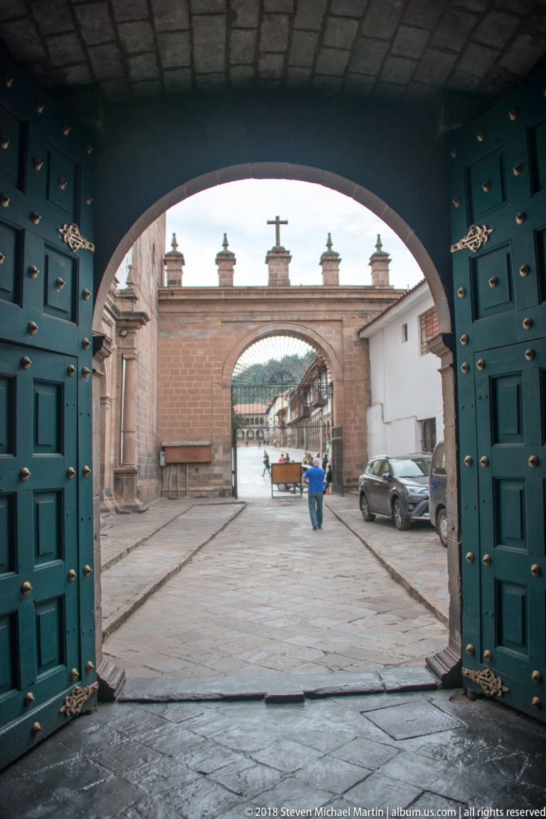 Open studded wooden doors frame a stone courtyard and a second arched gateway topped by a cross.