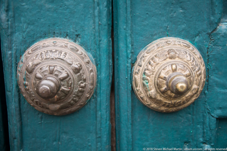 Two embossed circular metal door bosses with raised knobs mounted on a weathered turquoise wooden double door.
