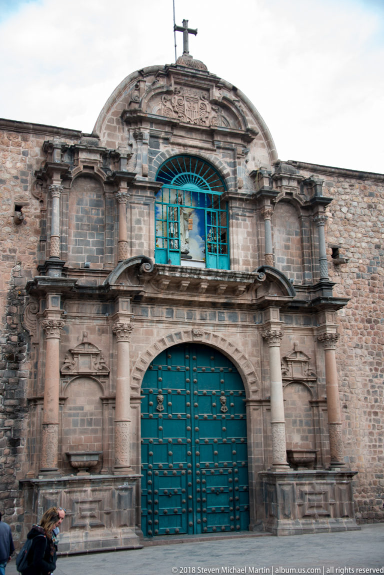 Ornate carved stone church portal with columns, niches, arched upper window and turquoise double doors.