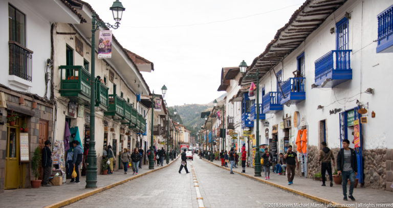 Straight Cusco street with pedestrians, shops and white buildings carrying green and blue wooden balconies.