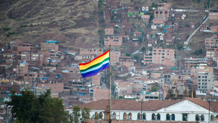 Seven-band rainbow flag flying above Cusco rooftops with dense brick hillside neighborhoods behind.