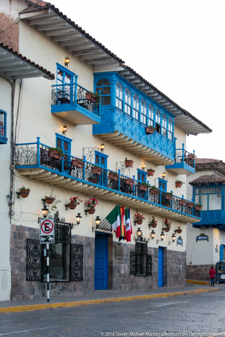 White colonial-style building with ornate bright blue wooden balconies, flower boxes, blue doors and a dark stone base.