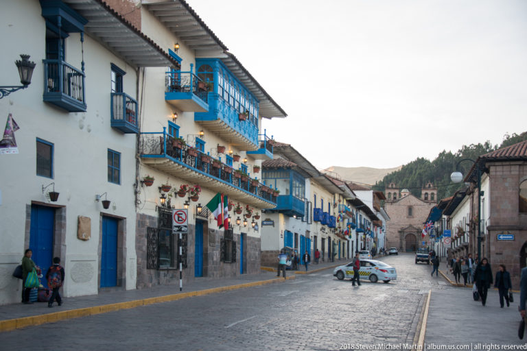 Cobbled Cusco street lined with white buildings and bright blue wooden balconies, with pedestrians and a church ahead.