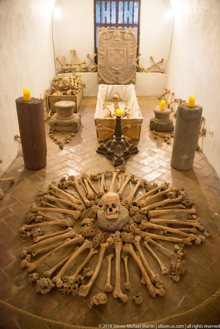 Human bones arranged in a radial floor pattern inside a whitewashed crypt with candles and funerary objects.