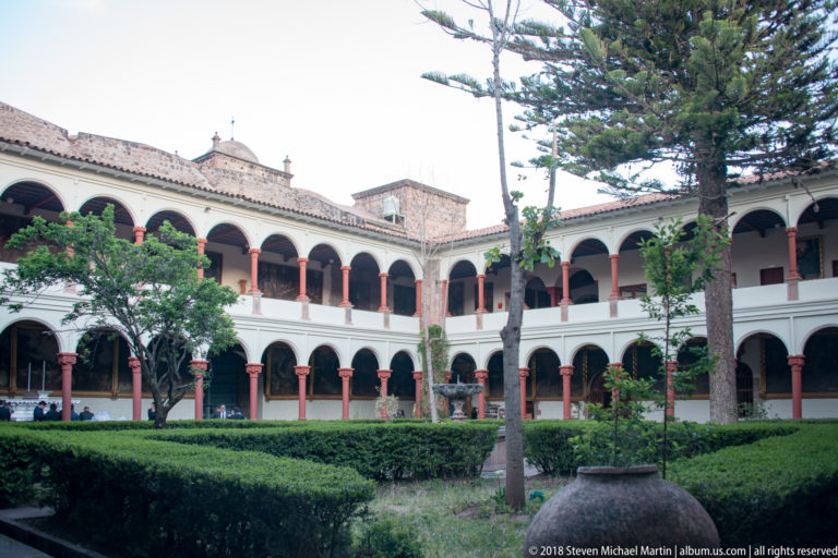 Tall conifer and clipped hedges inside a two-story cloister lined with white arches and red columns.