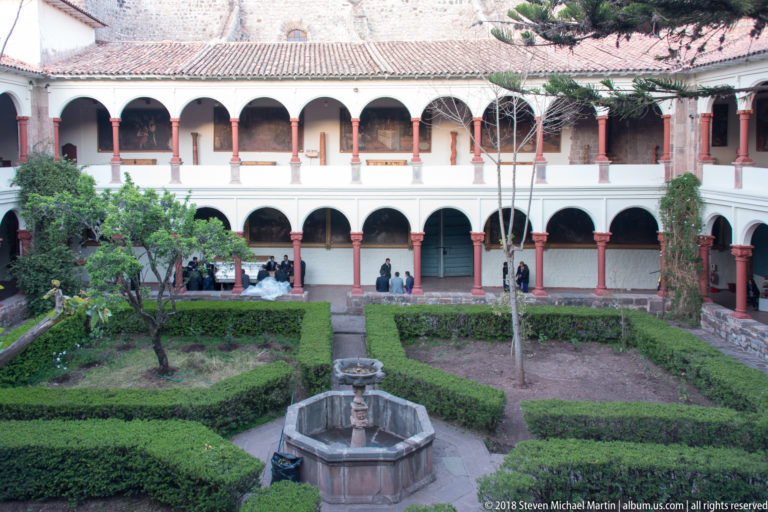 Geometric hedge garden and stone fountain enclosed by a two-story white cloister with red columns.