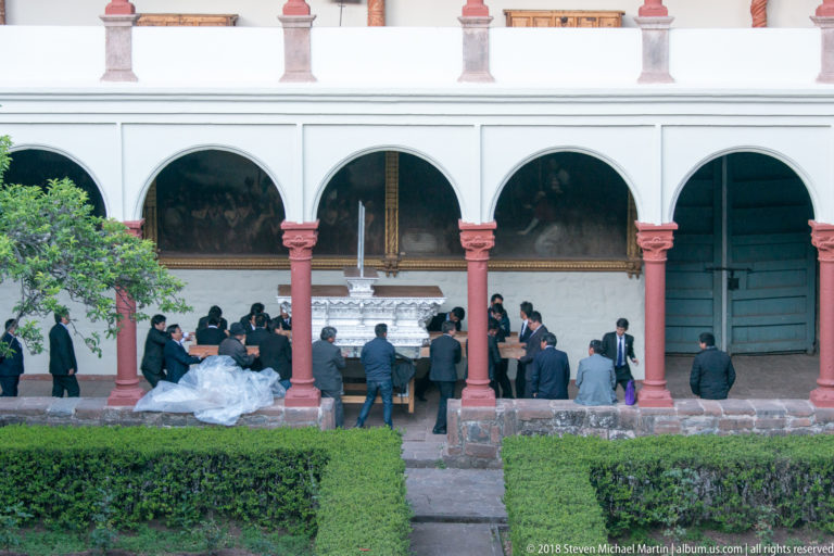Guests in formal clothing gather under a cloister arcade beside a person in a white dress, with hedges in front.