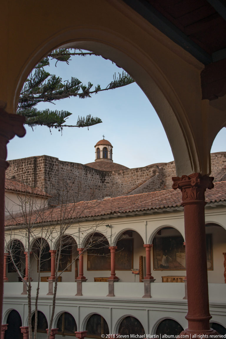 White two-story cloister with red stone columns, repeated arches, tiled roof and small cupola above.