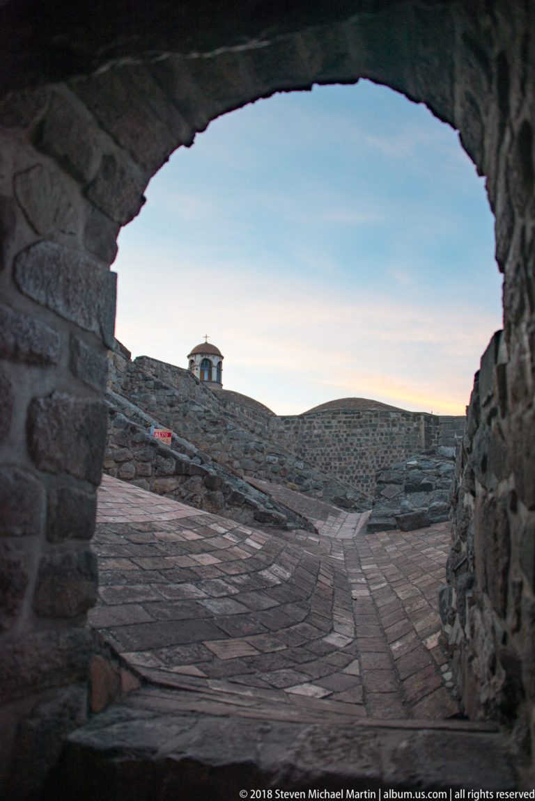 Rough stone arch framing a brick-paved passage between curved masonry walls with a small cupola beyond.