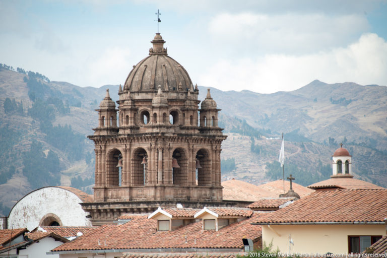 Large ribbed church dome with arched drum, corner turrets, cross, and red tile roofs against mountains.