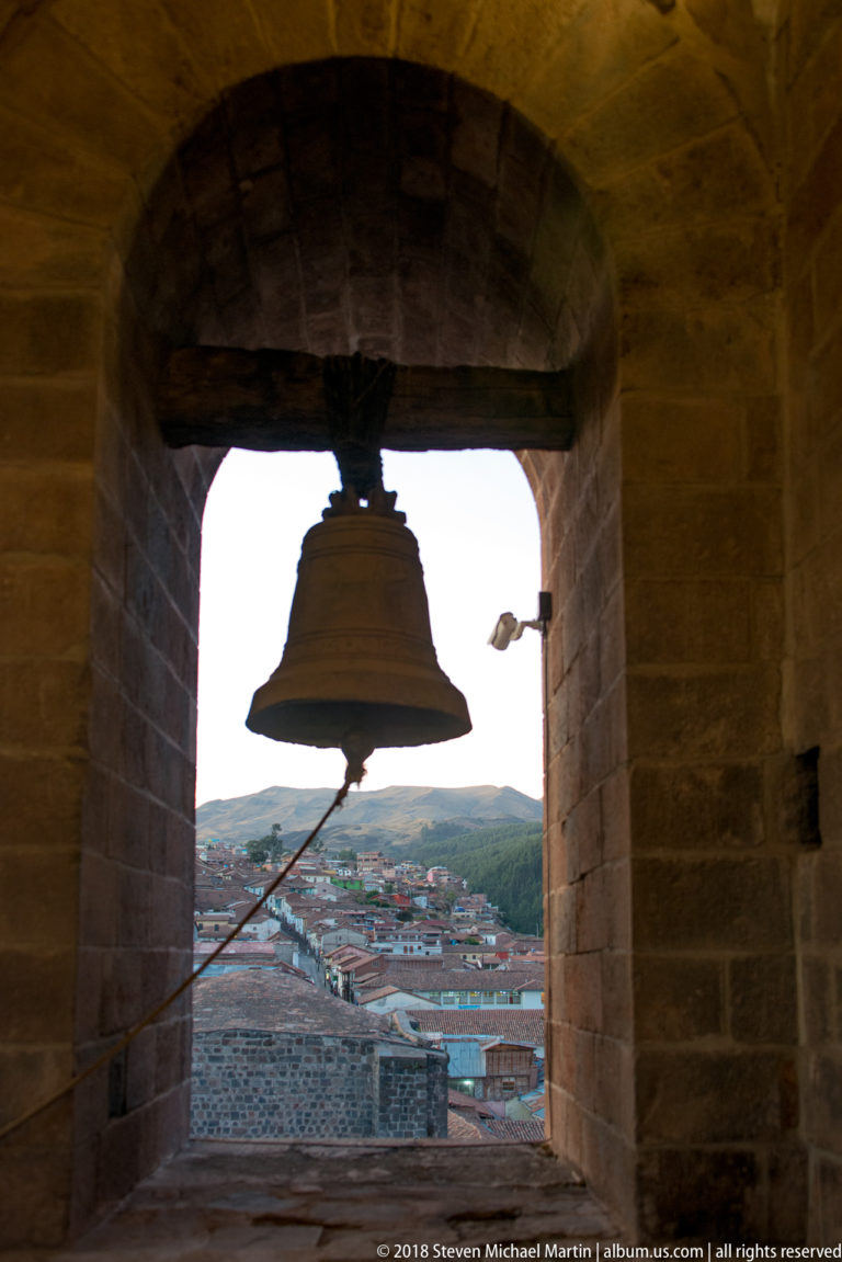 Silhouetted bell and diagonal rope inside a stone tower opening, with Cusco rooftops and hills beyond.