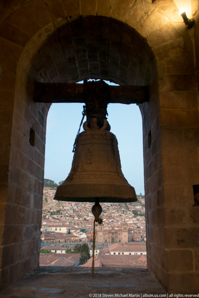 Bronze bell silhouetted in a stone belfry opening with Cusco rooftops visible through the arch behind it.