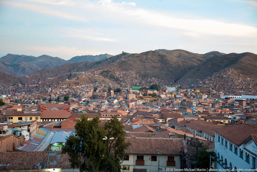 Elevated view over Cusco rooftops and hillside neighborhoods toward rounded Andean mountain ridges.