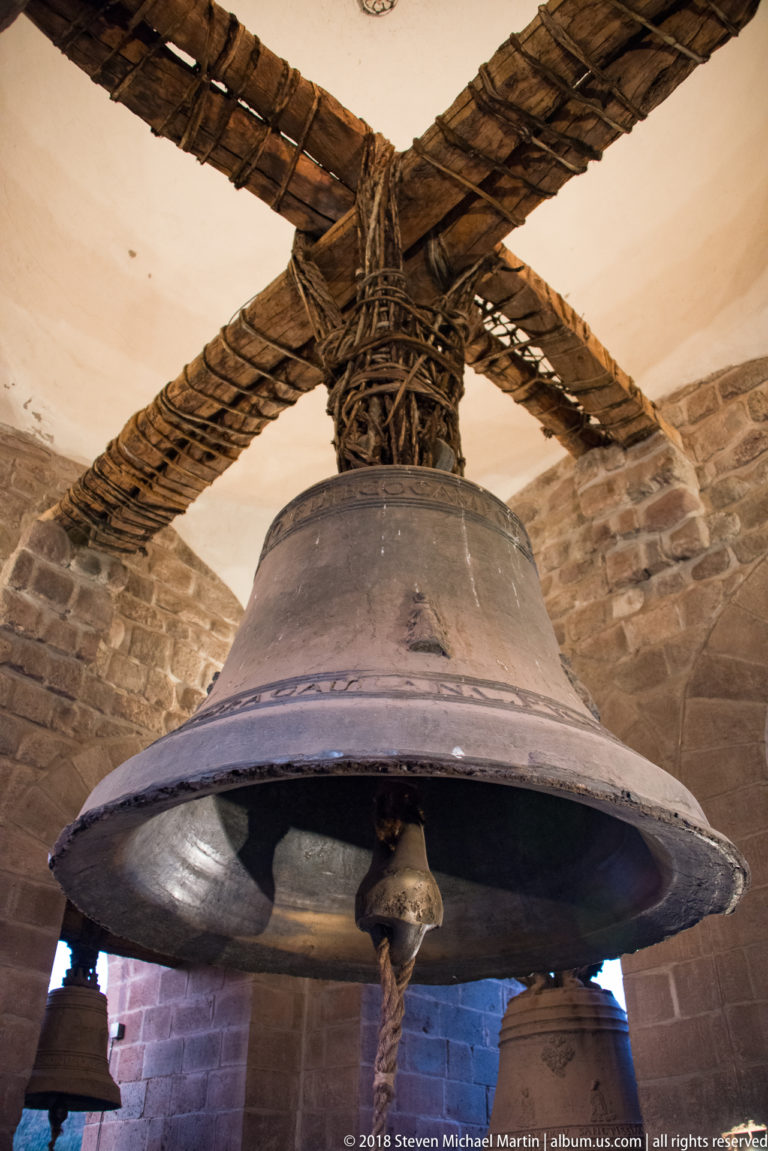 Large bronze bell hanging below rope-lashed wooden crossbeams inside a stone bell tower.