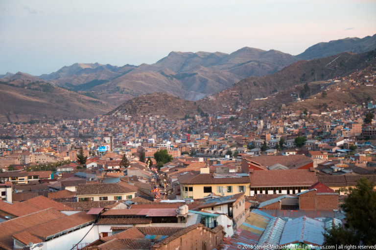 Wide Cusco cityscape filling a mountain basin, with red roofs, brick neighborhoods and dry Andean ridges beyond.