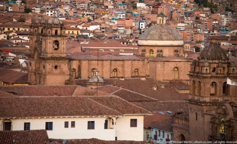 Multiple stone church towers and a dome rise above red-tiled roofs with a densely built hillside behind.