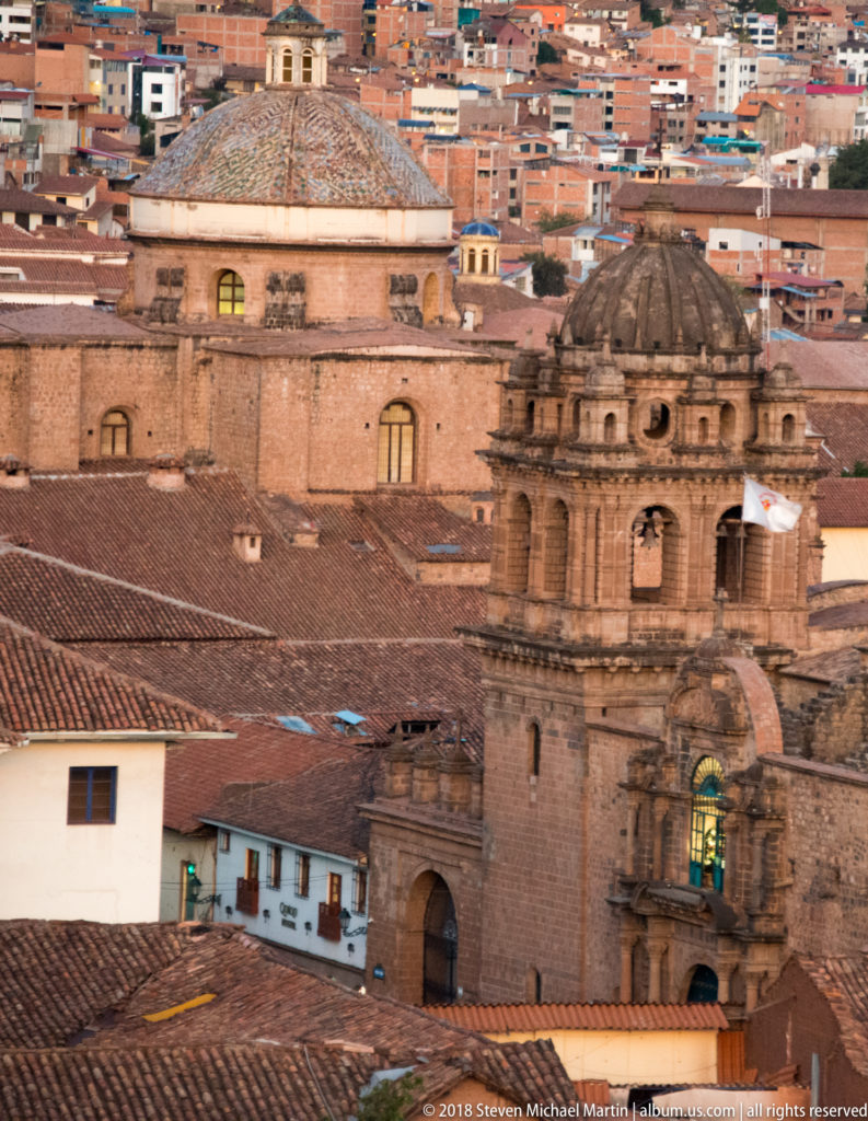 Stone church tower, dome and red-tiled roofs layered against densely built hillsides in Cusco.