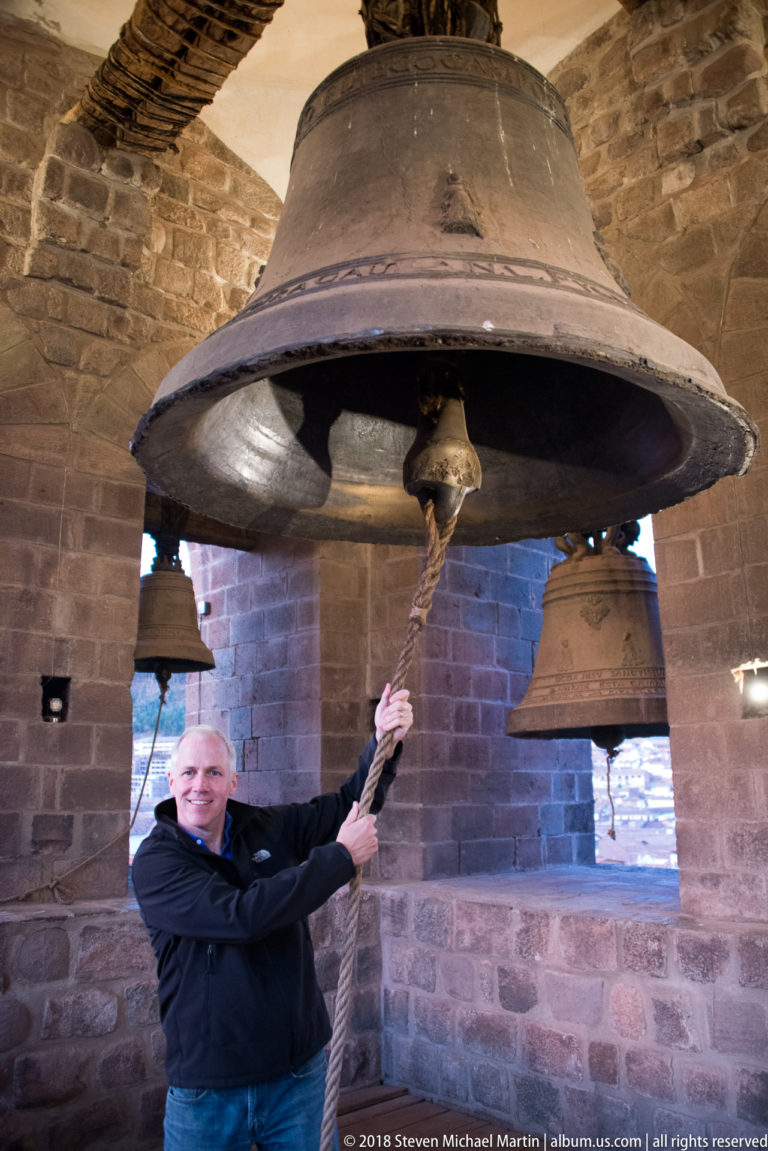 Large bronze bell suspended from timber beams in a stone tower as a man pulls its rope below.