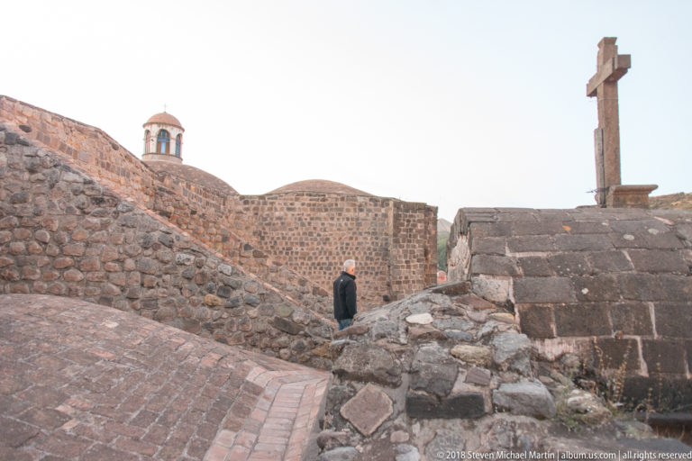Stone walls and brick paths curve around a stepped platform topped by a tall stone cross, with one person walking nearby.