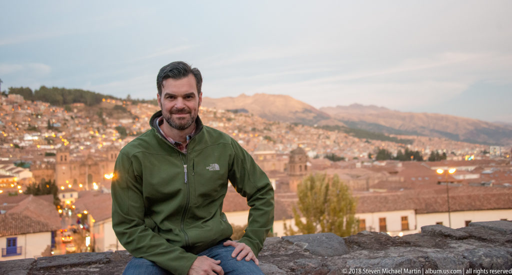 Seated man in a green jacket at a stone overlook with Cusco and Andean mountains spread behind him.