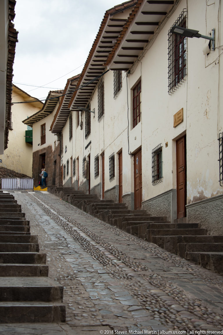 Steep cobblestone Cusco street with white houses, barred windows, wooden doors, and stone steps.