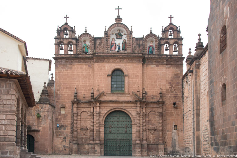 Stone church façade with green door, central window, bells, crosses, and painted religious niches along the roofline.