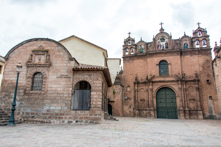 Small stone chapel beside a larger decorated church façade with bells, niches, and crosses.