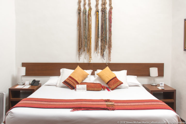 White bed with red woven runner, patterned cushions, wooden headboard, bedside lamps, and long fiber wall hangings.