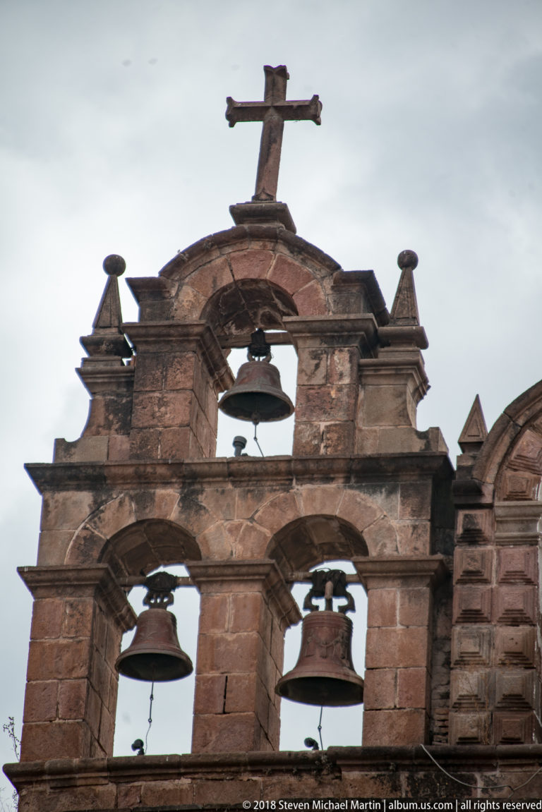 Frontal stone bell gable with two lower bells, one upper bell, finials, and summit cross.