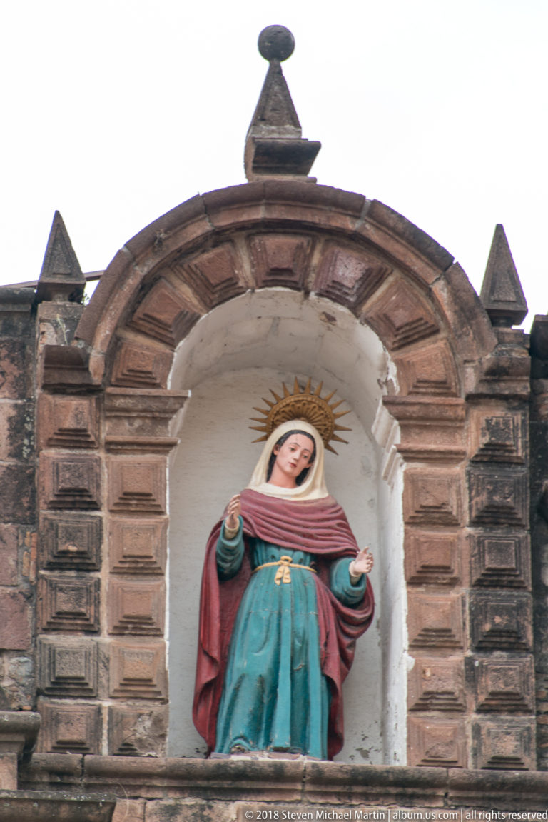 Polychrome haloed female religious figure in blue-green and rose garments within an arched stone niche.