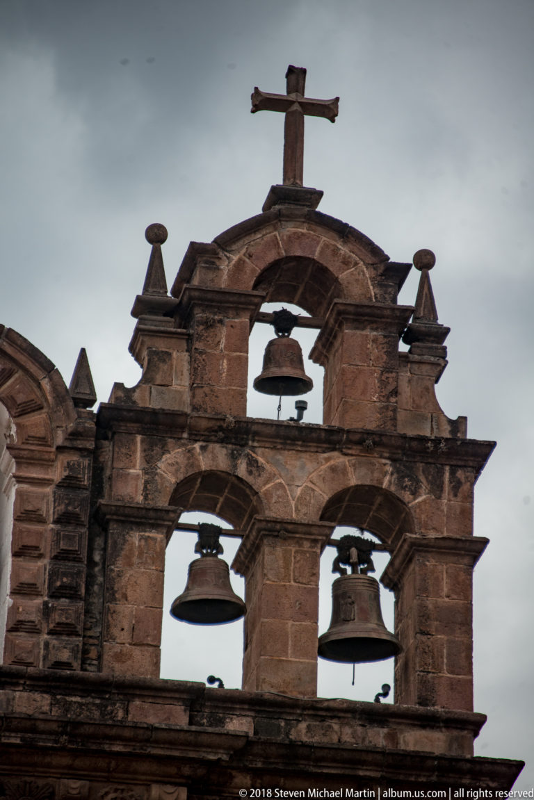 Weathered stone bell gable with three bells, corner finials, and a cross at the top.
