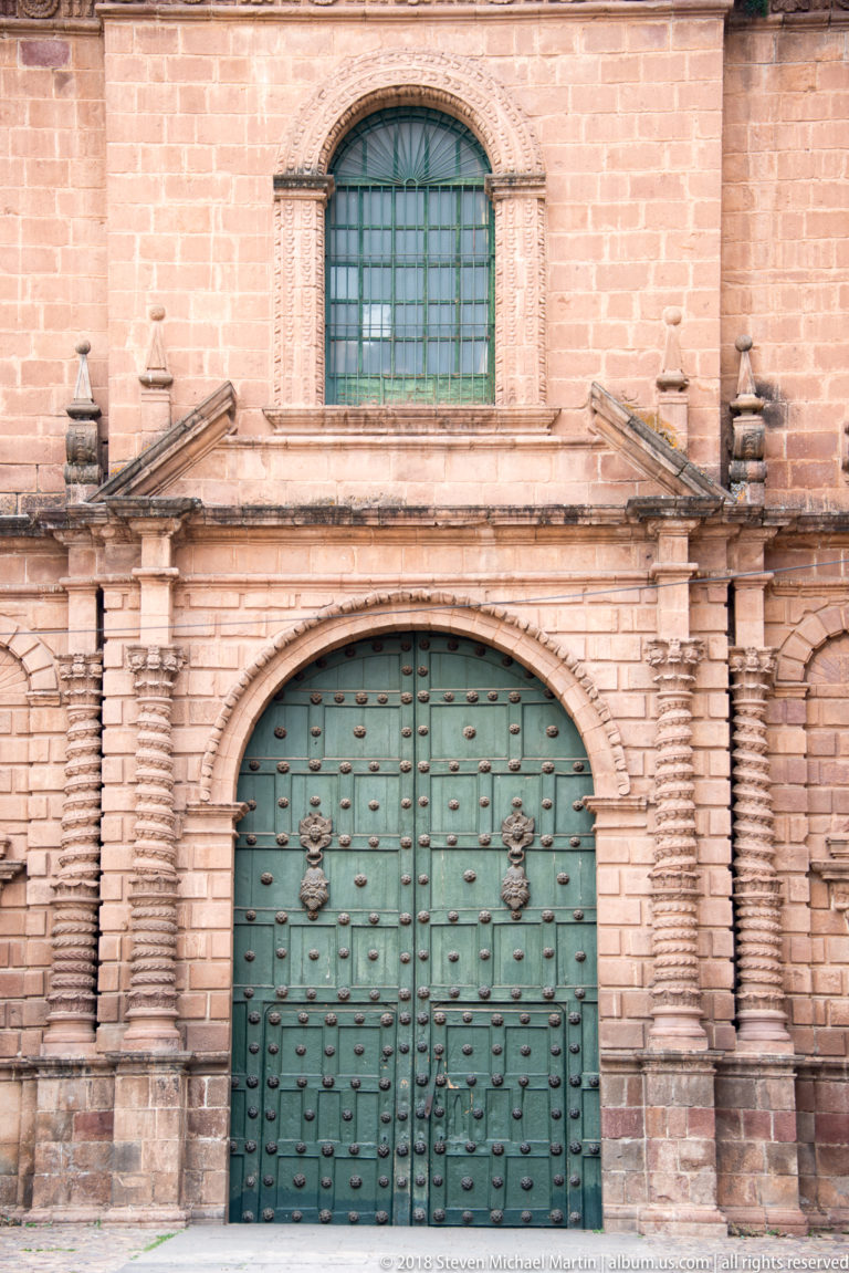 Green studded church doors beneath a large barred window in a symmetrical stone façade.