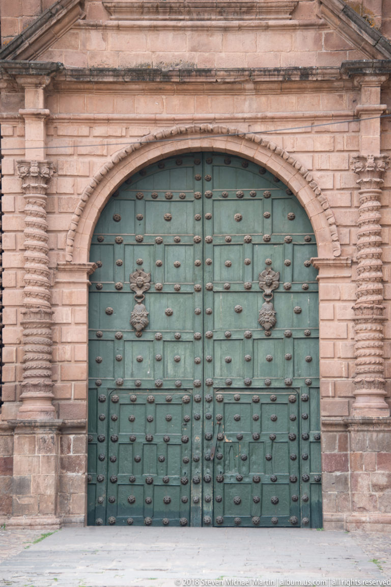 Wide green studded double doors with two heraldic plaques in a rusticated stone church façade.