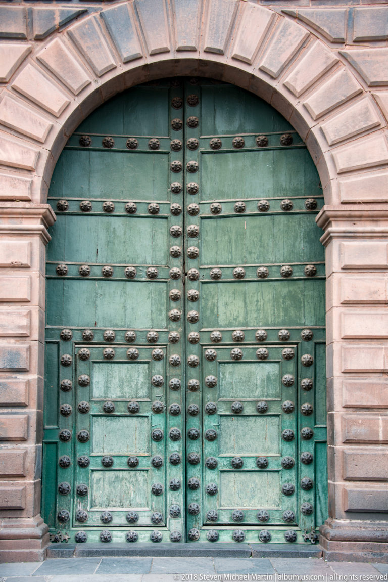 Weathered green double doors densely covered with rows of dark metal bosses beneath a stone arch.