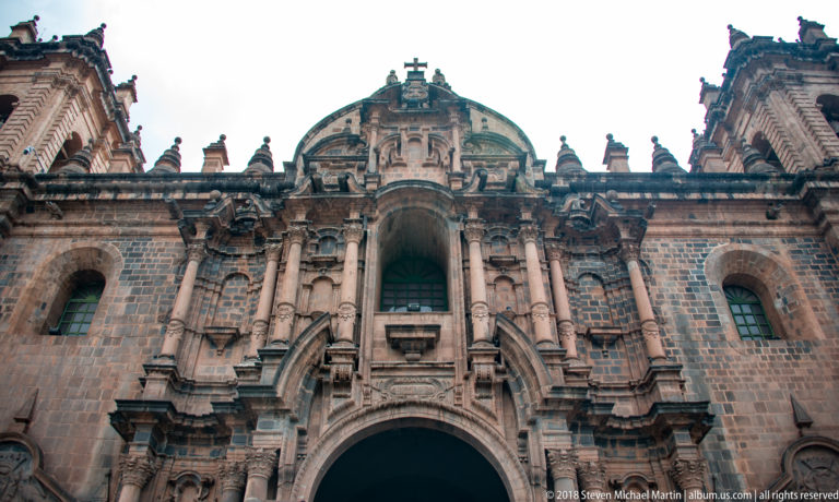Upward view of ornate baroque stone church façade with columns, niches, and a large arched window.