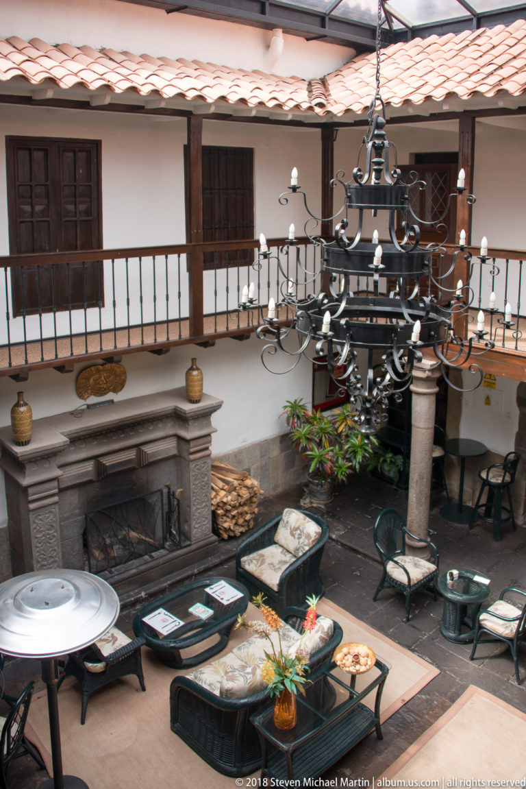 Furnished courtyard with large hanging metal chandelier, wooden balcony, fireplace, tables, and cushioned seating.