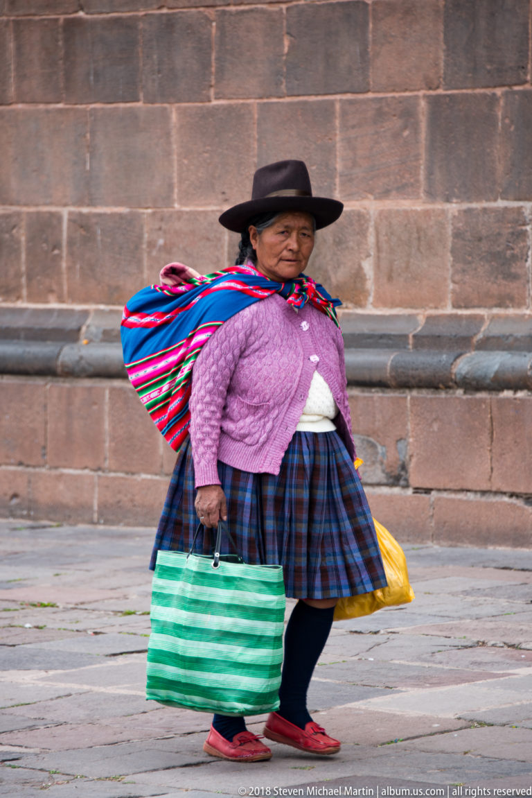 Andean woman in a dark hat and layered skirt carrying shopping bags and a colorful woven bundle.