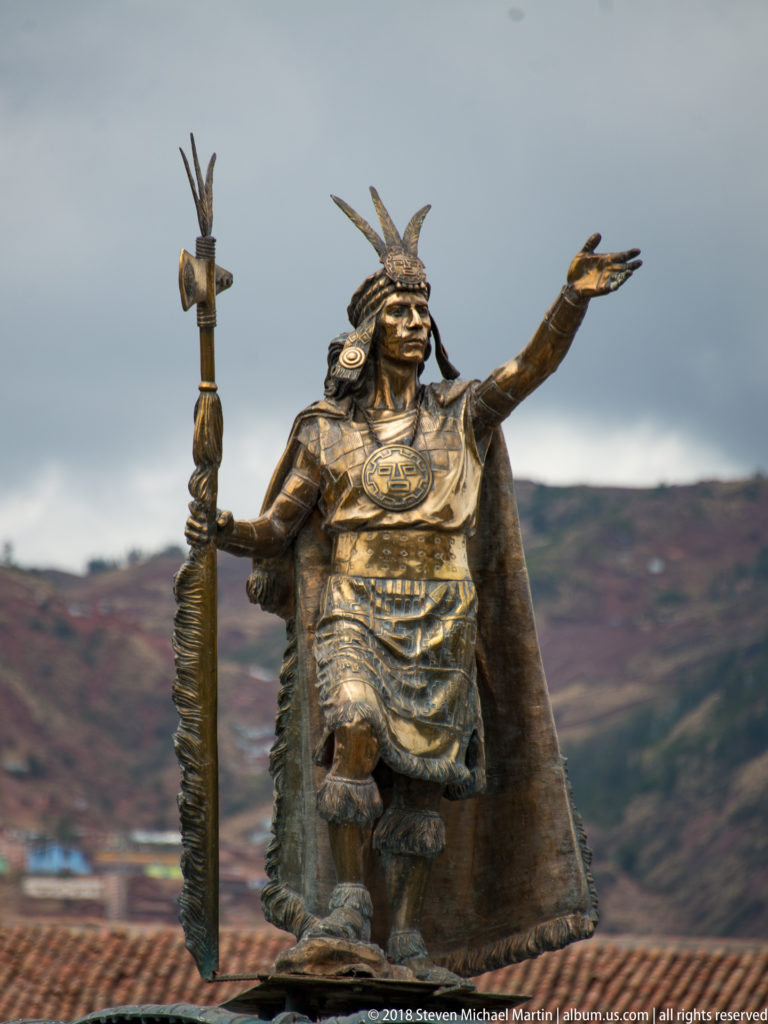 Close view of Inca ruler statue with feathered headdress, staff, patterned tunic, and extended hand.