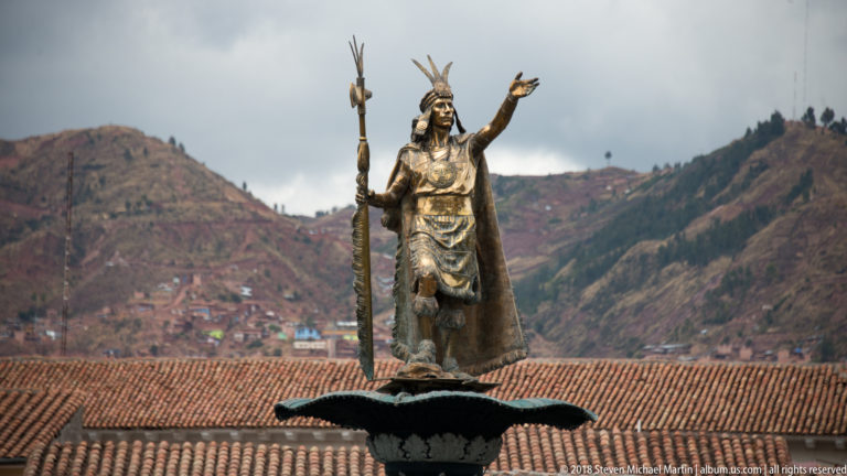 Bronze-colored Inca ruler statue holding a staff above red tile roofs and mountain slopes.