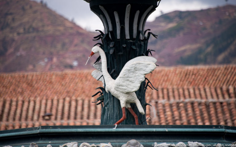 White long-legged bird sculpture with spread wings attached to the dark fountain column.