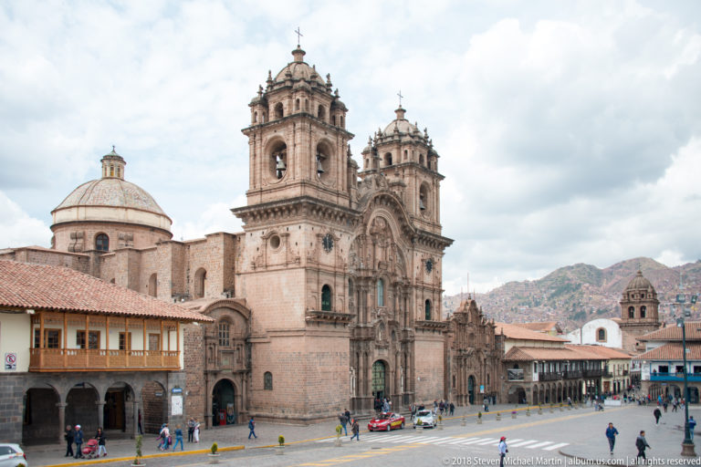 Twin-towered stone church with ornate central façade and large dome beside Cusco plaza buildings.