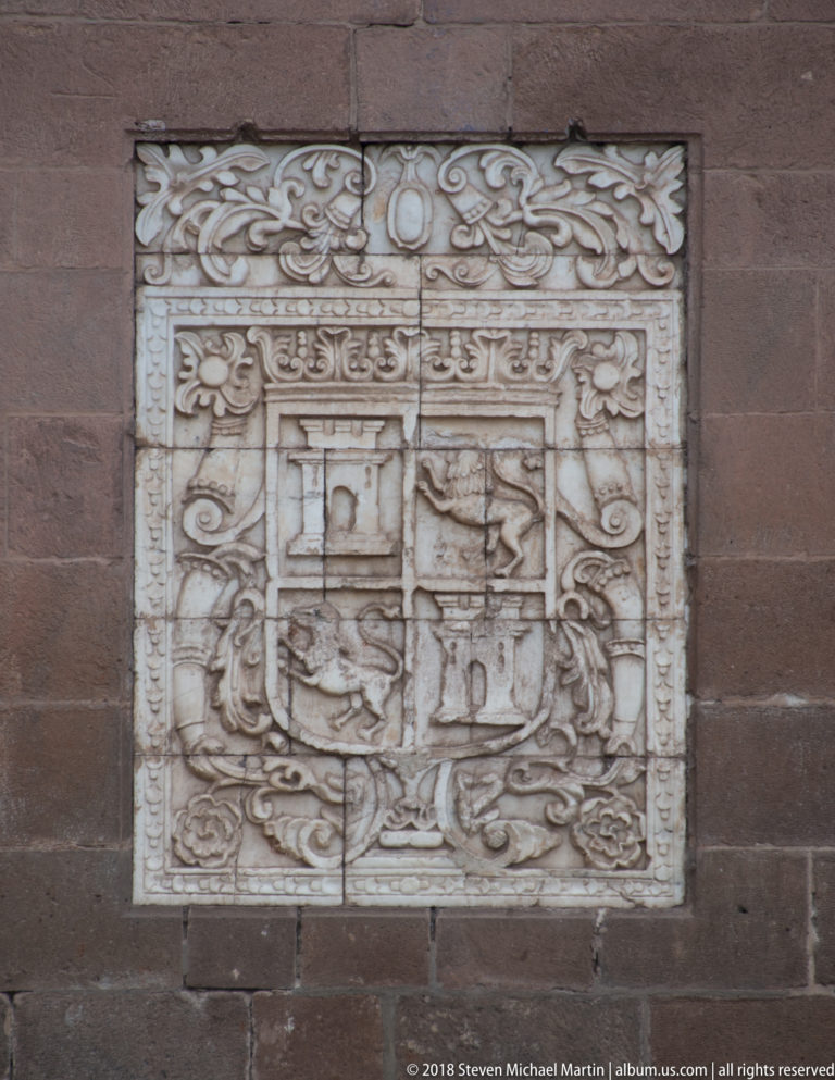 Carved heraldic shield with scrolling ornament inset into a dark stone wall.