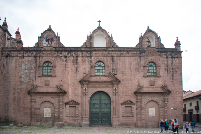 Wide stone church façade with tall central door, two smaller side portals, upper windows, and roofline ornament.