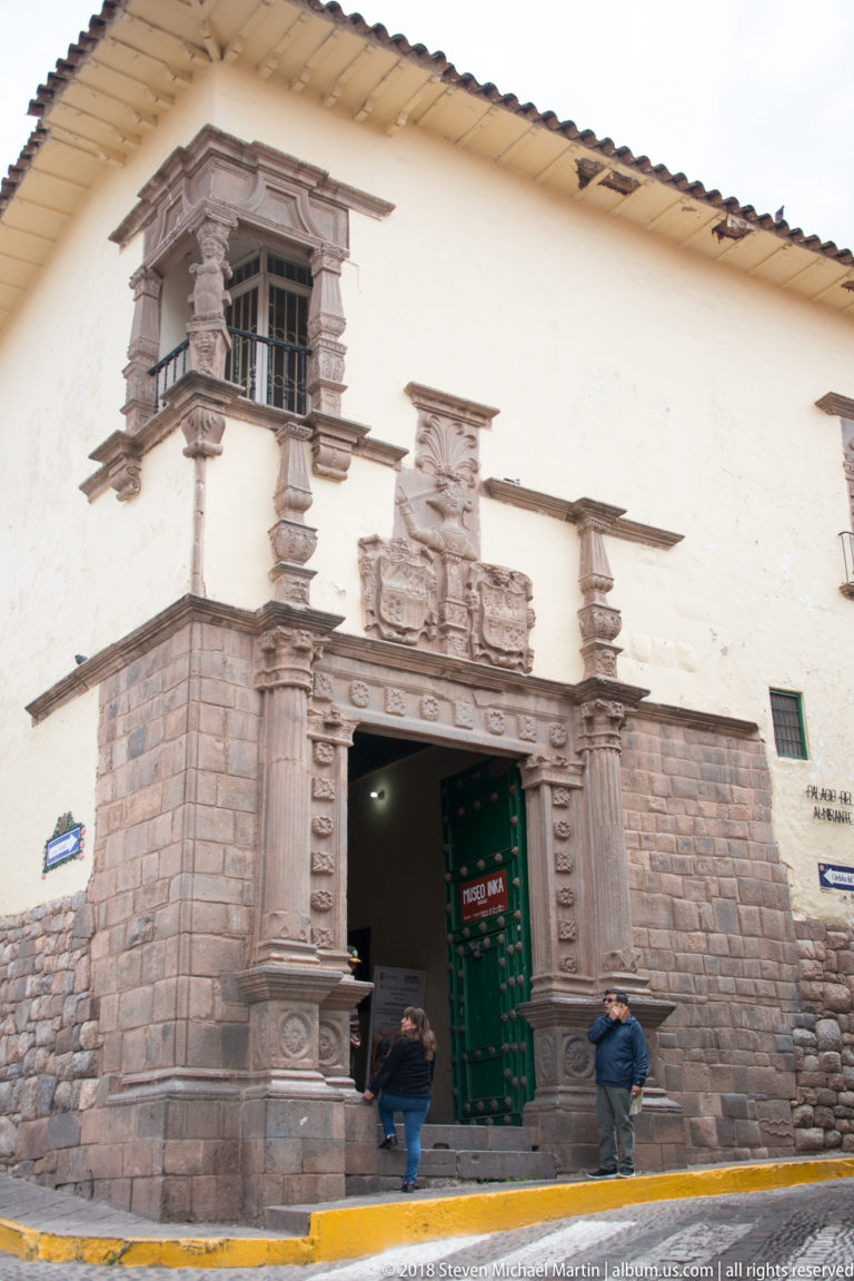 White historic façade with large carved stone entrance, heraldic relief, and projecting enclosed window.