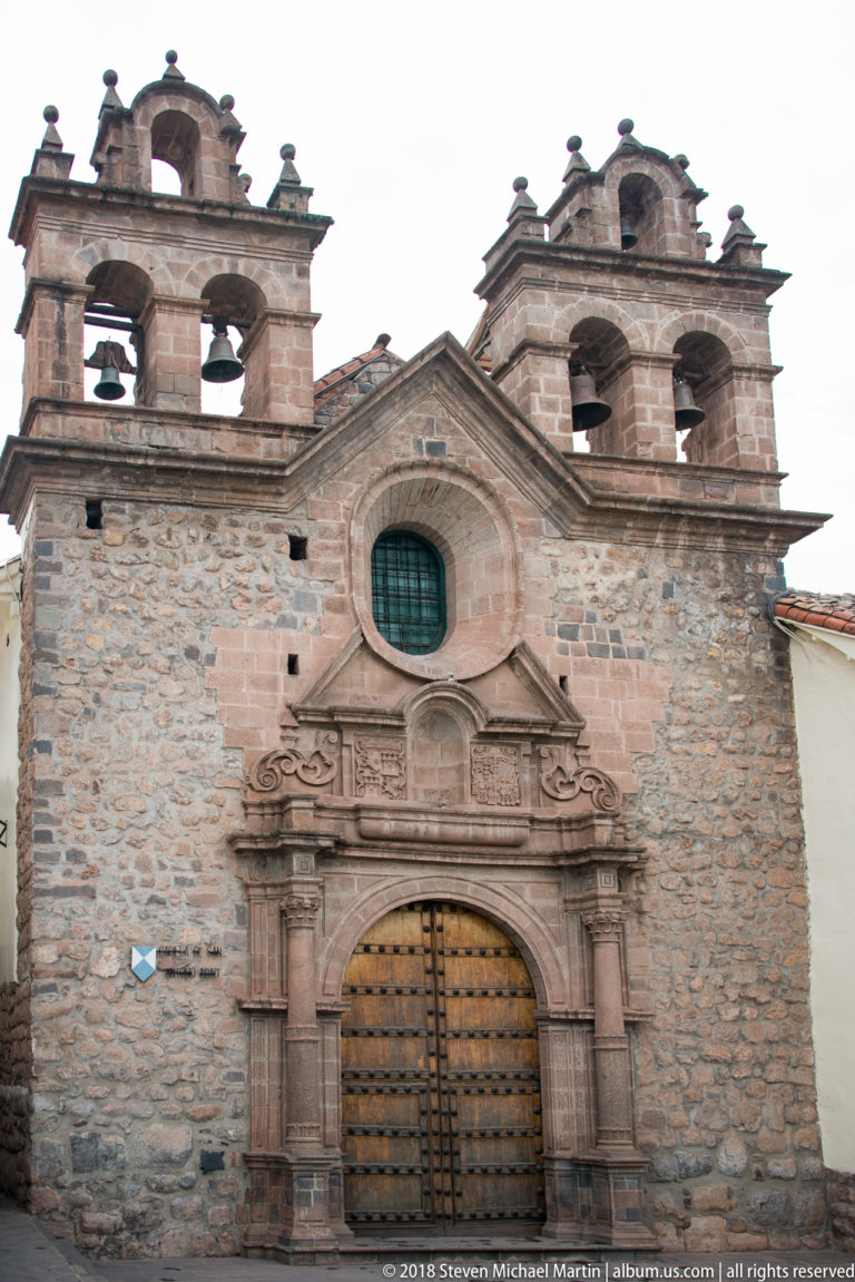 Stone church façade with twin bell gables, oval window, and large studded wooden door.