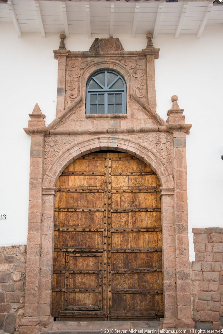 Large studded wooden double door framed by carved stonework and a small arched window above.