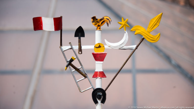 Colorful metal weather vane with rooster, crescent, star, ladder, shovel, pickaxe, and yellow ornament.