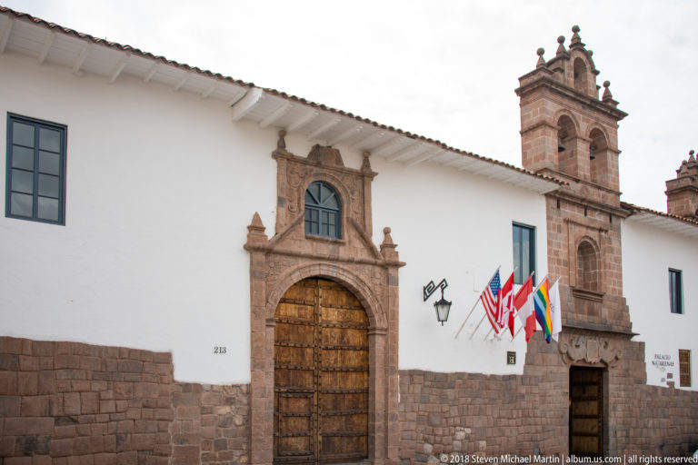 White historic façade with carved stone doorway, wooden door, flags, and a tall bell tower.