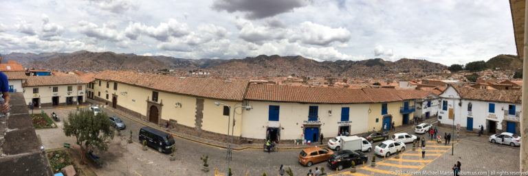 Elevated Cusco street view with tiled roofs, vehicles, dense hillside development, and rugged mountains.