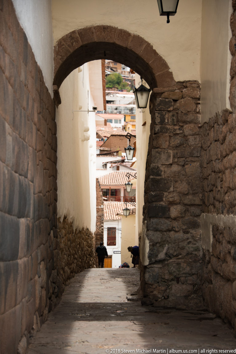 Stone-lined passage under a broad arch, opening toward tiled roofs and city buildings beyond.